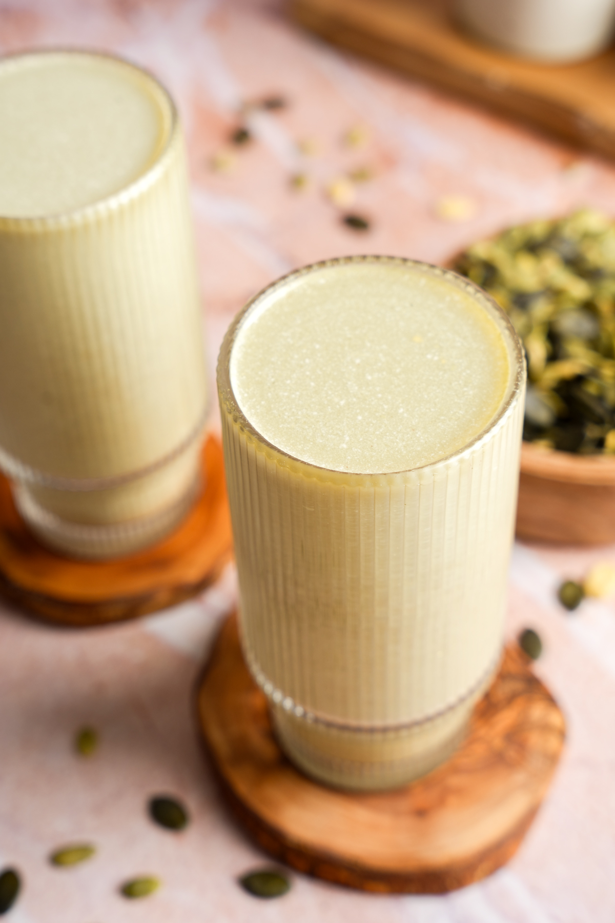 Close-up of a glass filled with creamy homemade pumpkin seed milk, placed on a wooden coaster with pumpkin seeds scattered across a pink textured surface.