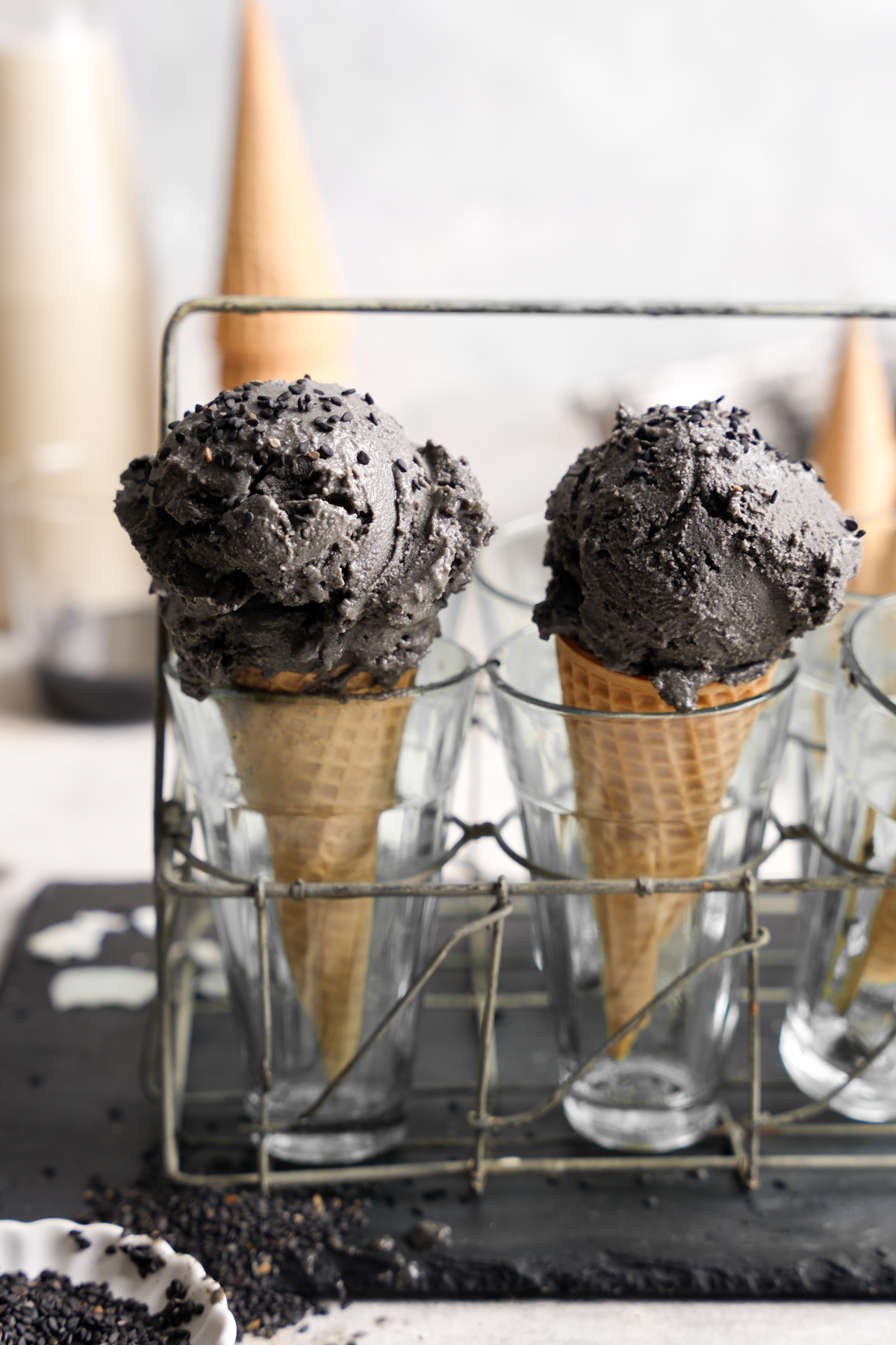 Two waffle cones filled with scoops of rich black sesame ice cream, displayed upright in clear glasses within a metal wire rack, with black sesame seeds scattered below.