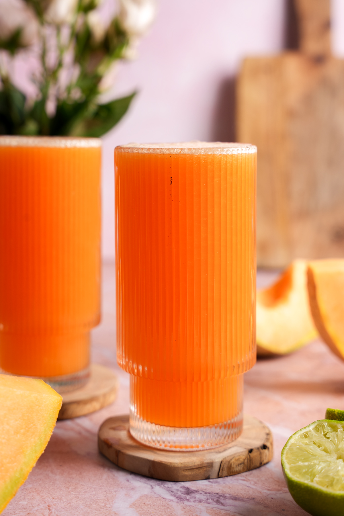 Homemade cold-pressed cantaloupe juice served in textured glasses on wooden coasters, surrounded by fresh cantaloupe slices and halved limes on a light pink marble surface
