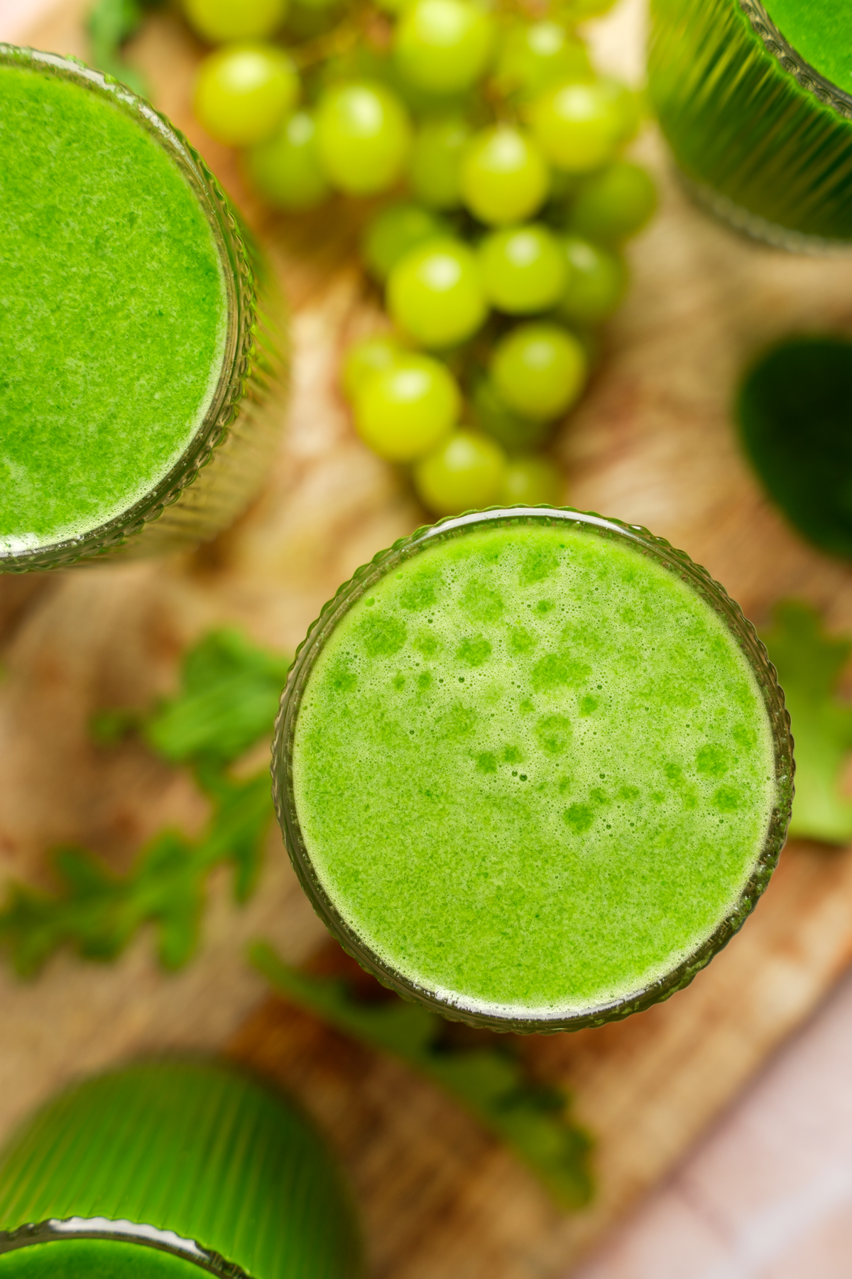 Top-down view of vibrant homemade green juice in clear glasses, surrounded by fresh green grapes, arugula, and spinach — refreshing detox juice made with kale, cucumbers, and green apples.