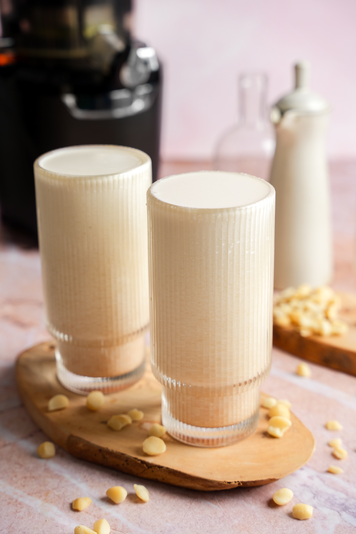 Two glasses of homemade macadamia nut milk on a wooden board with scattered macadamia nuts, made using a slow juicer, on a pink marble background.