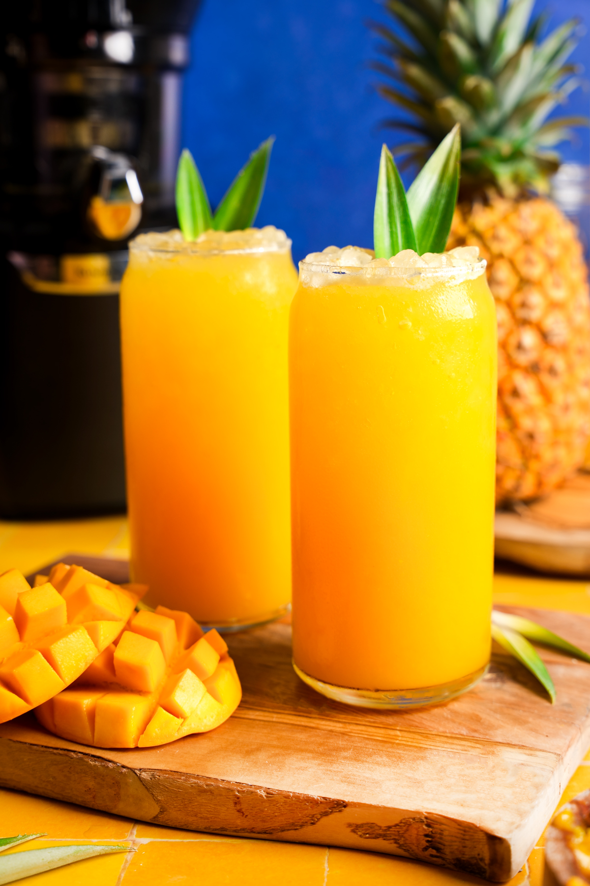Two tall glasses of homemade mango pineapple juice topped with crushed ice and garnished with pineapple leaves, served on a wooden board with fresh mango cubes, a whole pineapple, and a juicer in the background for a vibrant tropical drink presentation.