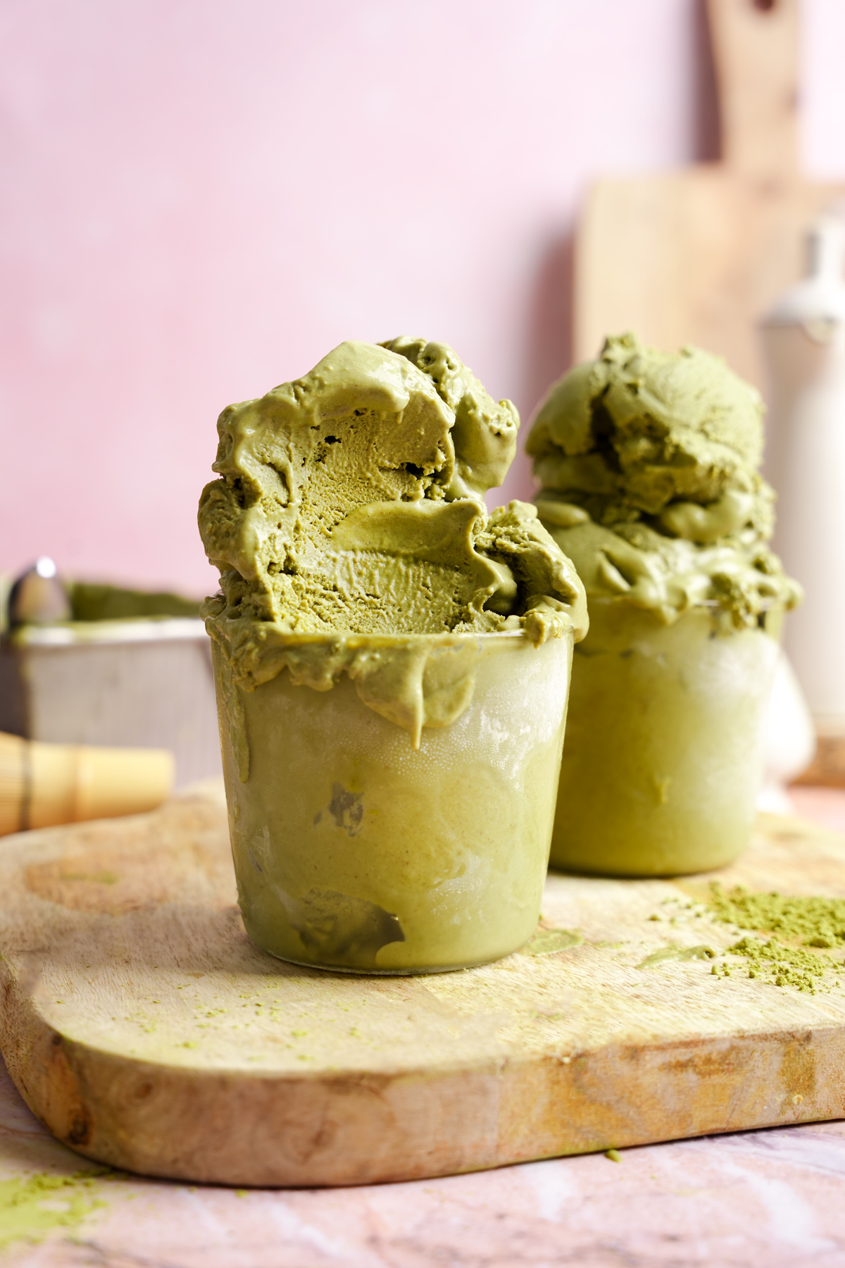 Creamy vegan matcha ice cream served in frosty glass cups on a wooden board, with a pink background and matcha powder sprinkled nearby.
