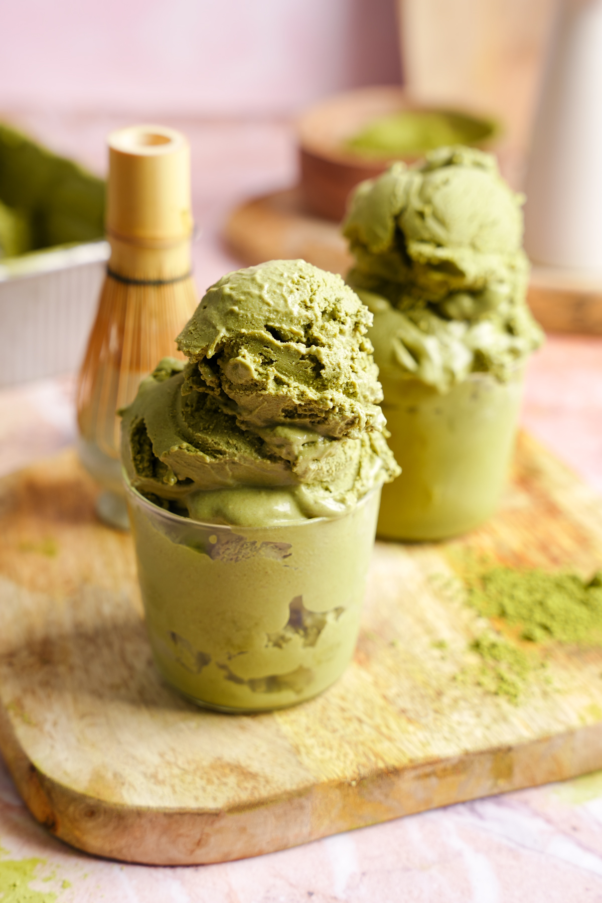 Two glass cups filled with creamy homemade vegan matcha ice cream, placed on a wooden board with a bamboo matcha whisk and powdered green tea in the background.