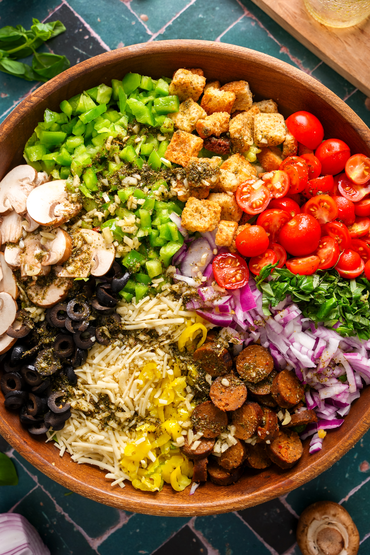Overhead view of a deconstructed vegan pizza salad in a wooden bowl with neatly arranged toppings including cherry tomatoes, croutons, green bell peppers, mushrooms, black olives, shredded vegan cheese, banana peppers, vegan sausage, red onions, and fresh basil