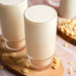 Glass of creamy homemade macadamia nut milk on a wooden serving board, surrounded by chopped raw macadamia nuts with a soft pink backdrop.
