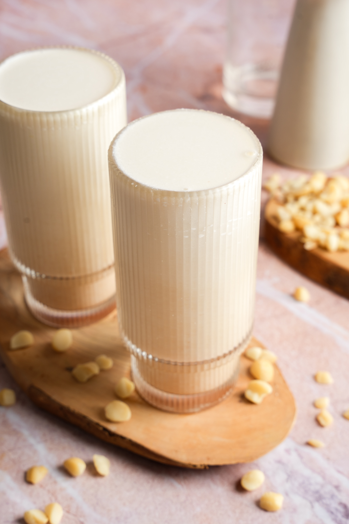 Two tall glasses of creamy homemade macadamia nut milk on a wooden serving board, surrounded by scattered raw macadamia nuts, with a soft pink marble background and minimalist kitchen elements in the distance.