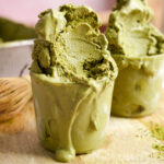 Vegan matcha ice cream overflowing in glass cups, rich and creamy with a vibrant green hue, melting over a wooden board with bamboo whisk and matcha powder in the background.