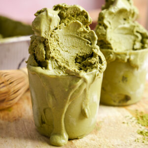 Vegan matcha ice cream overflowing in glass cups, rich and creamy with a vibrant green hue, melting over a wooden board with bamboo whisk and matcha powder in the background.
