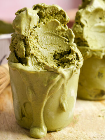 Vegan matcha ice cream overflowing in glass cups, rich and creamy with a vibrant green hue, melting over a wooden board with bamboo whisk and matcha powder in the background.