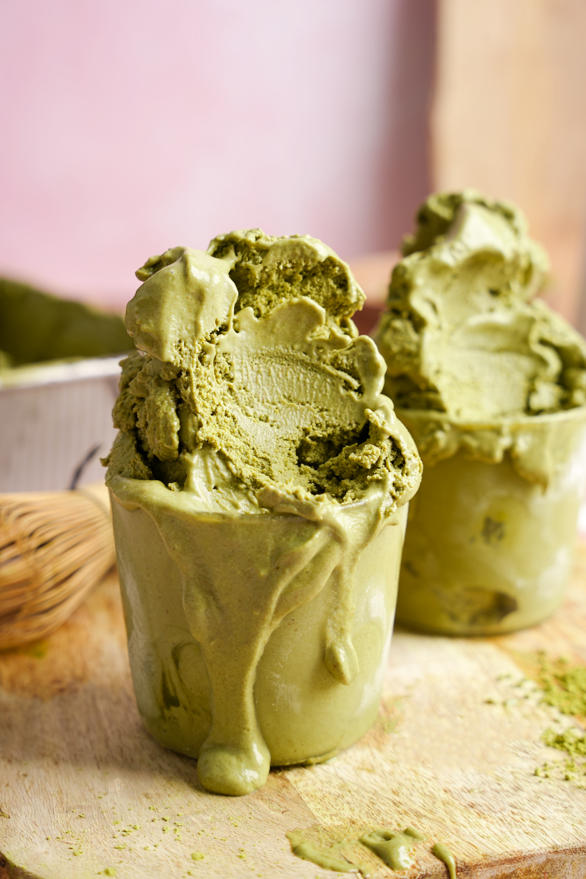 Close-up of creamy vegan matcha ice cream melting over the edge of a glass, with another serving in the background on a wooden surface, surrounded by green tea powder.