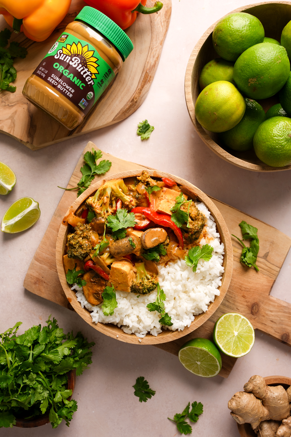 Nut-free vegan tofu Panang curry served with fluffy white rice in a wooden bowl, topped with fresh cilantro and surrounded by fresh limes, ginger, bell peppers, cilantro, and a jar of SunButter Organic Sunflower Seed Butter.