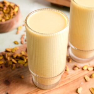 Tall glass of creamy pistachio nut milk on a wooden board, surrounded by chopped pistachios and a rustic bowl in the background, showcasing a smooth, rich texture and natural green hue.