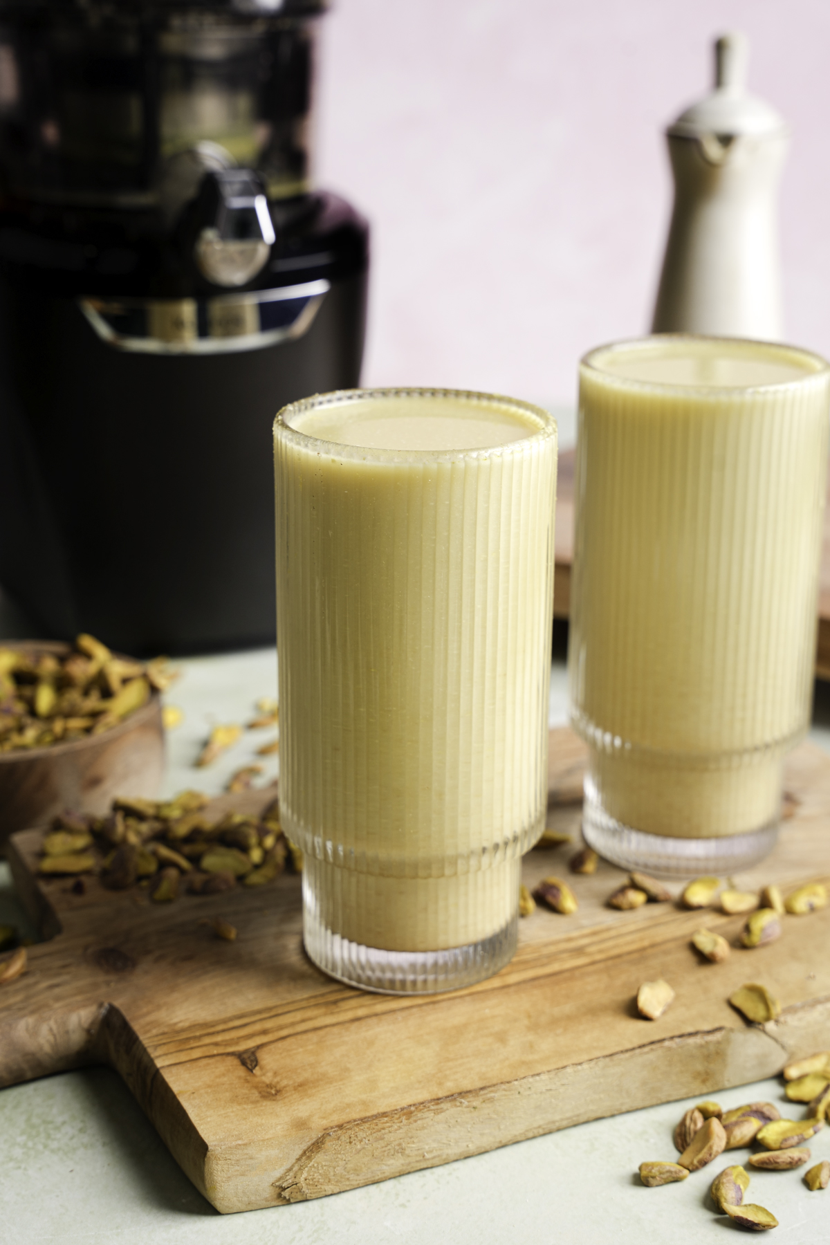 Two tall glasses filled with homemade pistachio nut milk on a wooden cutting board, surrounded by shelled pistachios and a juicer in the background.