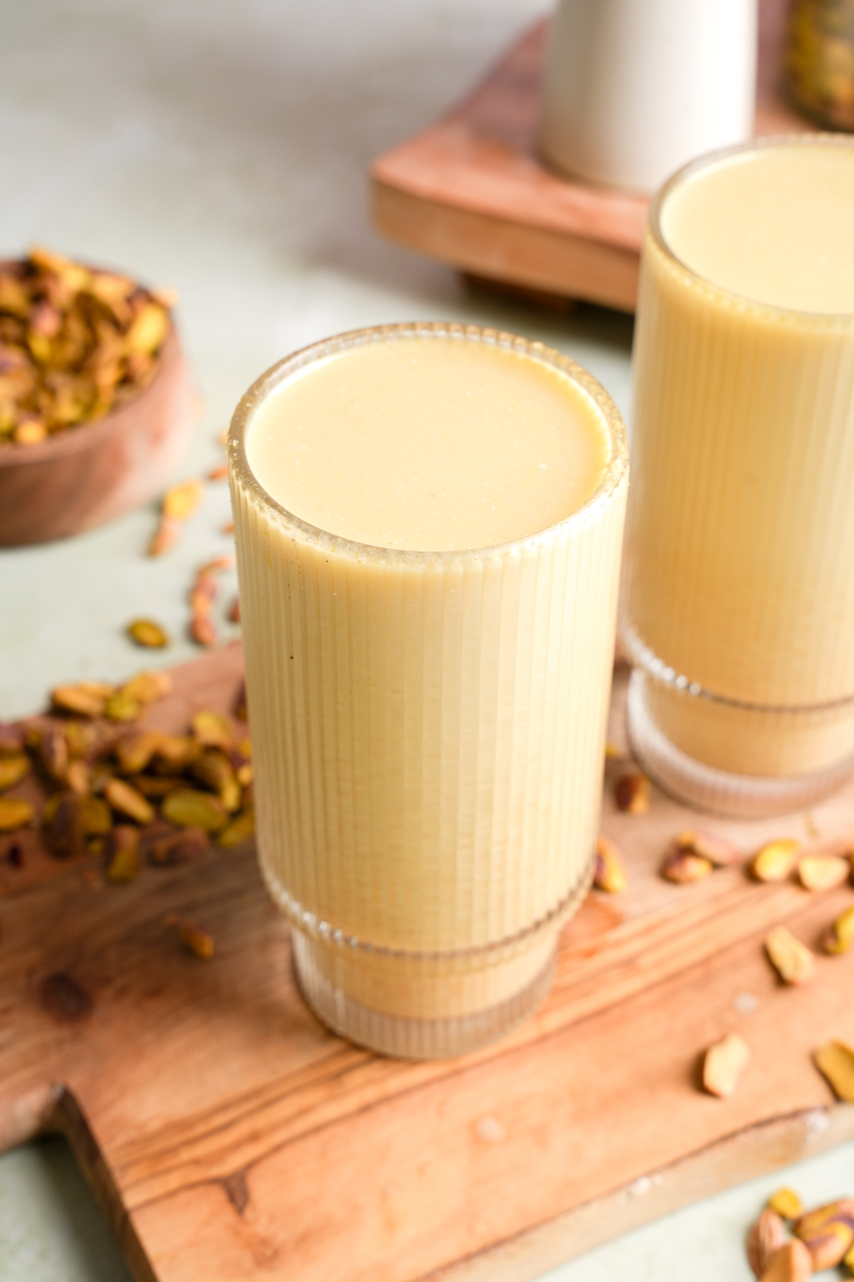 Close-up of a glass filled with creamy homemade pistachio nut milk, placed on a wooden board with scattered pistachios and a bowl of shelled pistachios in the background.