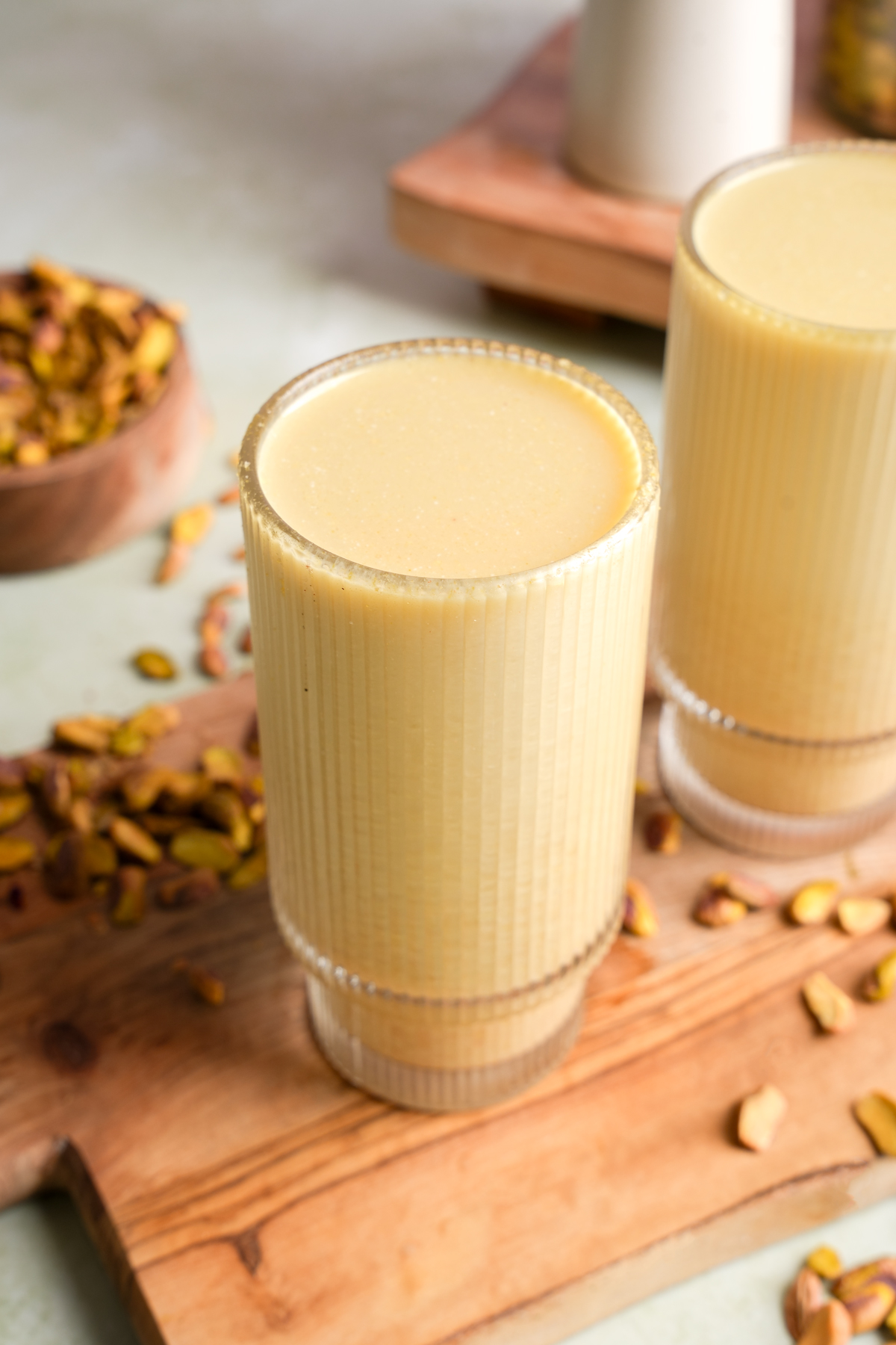 Glass of creamy homemade pistachio nut milk on a wooden board, surrounded by shelled pistachios and a second glass in the background.