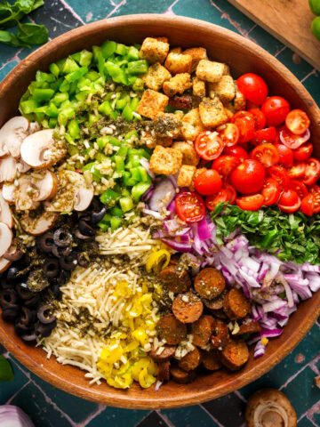 Overhead view of a deconstructed vegan pizza salad in a wooden bowl with neatly arranged rows of cherry tomatoes, croutons, green bell peppers, mushrooms, olives, vegan cheese, banana peppers, vegan sausage, red onion, and fresh basil.
