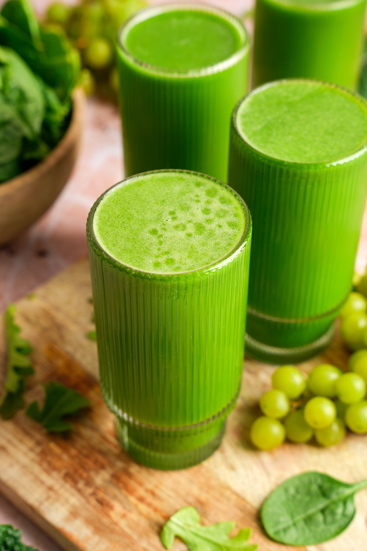 Vibrant green juice served in tall ribbed glasses on a wooden board, surrounded by fresh spinach, kale, arugula, and green grapes, highlighting a refreshing and nutrient-dense detox drink.