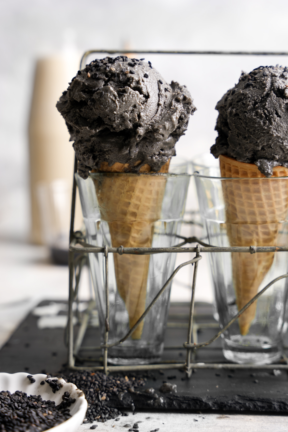 Creamy scoops of black sesame ice cream served in waffle cones, placed upright in clear glasses within a rustic wire holder, with black sesame seeds scattered on the surface below.