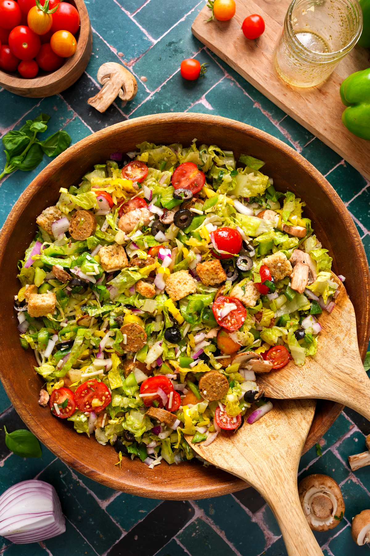 Chopped vegan pizza salad in a large wooden bowl with cherry tomatoes, black olives, green bell peppers, croutons, vegan sausage, mushrooms, red onions, and shredded lettuce, served with wooden salad utensils on a tiled surface