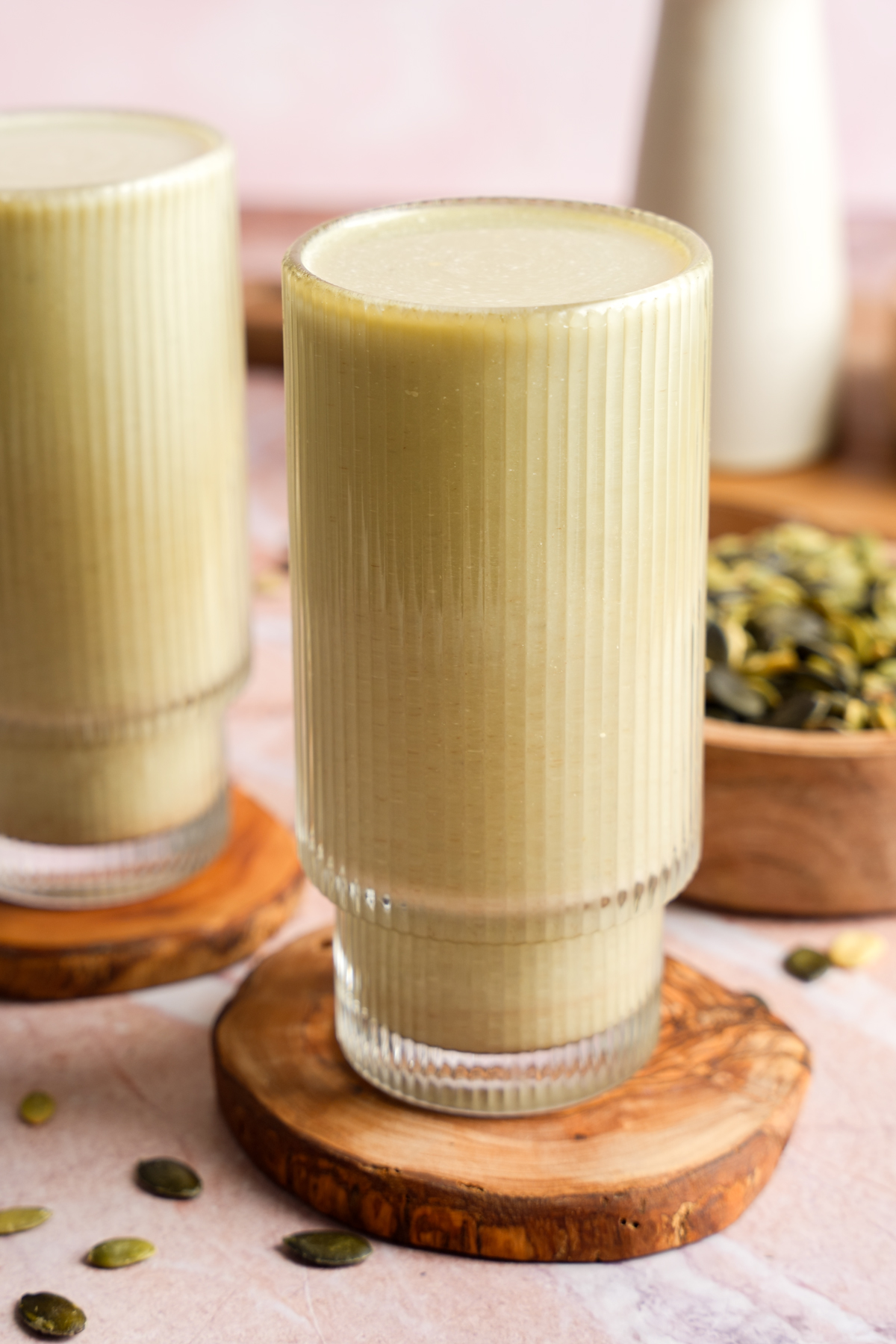 Glass of creamy vegan pumpkin seed milk on a wooden coaster, surrounded by scattered pumpkin seeds and a wooden bowl filled with more seeds in the background.