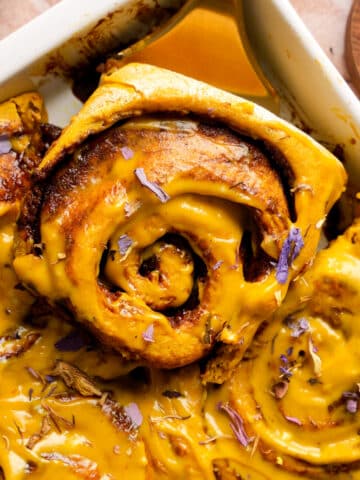 Close-up of a golden milk cinnamon roll being lifted from a baking dish, swirled with turmeric glaze and sprinkled with dried edible flower petals.