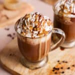 A glass mug of thick and creamy vegan hot chocolate topped with a generous swirl of dairy-free whipped cream, drizzled with rich chocolate sauce and sunbutter, set on a wooden board with scattered mini chocolate chips and a blurred second mug in the background.