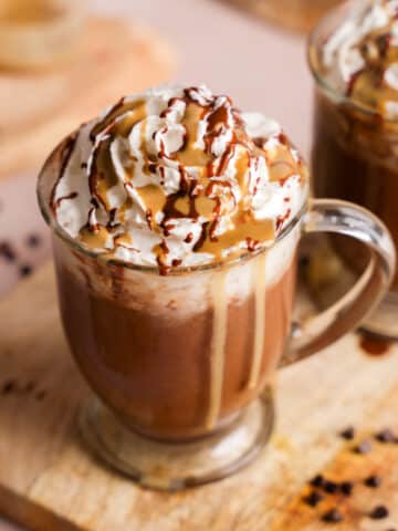 A glass mug of thick and creamy vegan hot chocolate topped with a generous swirl of dairy-free whipped cream, drizzled with rich chocolate sauce and sunbutter, set on a wooden board with scattered mini chocolate chips and a blurred second mug in the background.