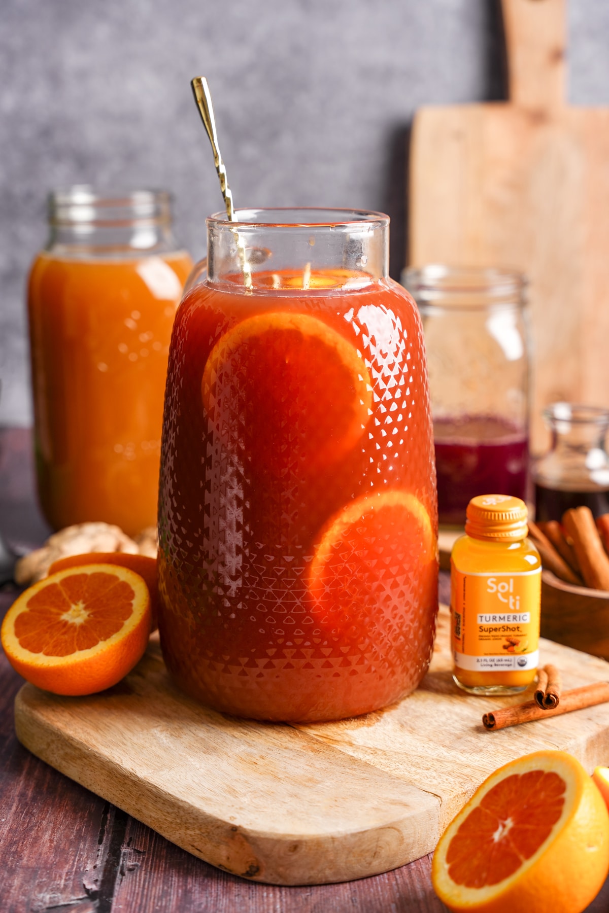 A glass pitcher filled with vibrant cranberry apple cider punch garnished with orange slices sits on a wooden board. Surrounding the pitcher are fresh oranges, cinnamon sticks, ginger, and a Sol-ti Turmeric SuperShot, creating a cozy and health-boosting autumn-inspired drink scene.