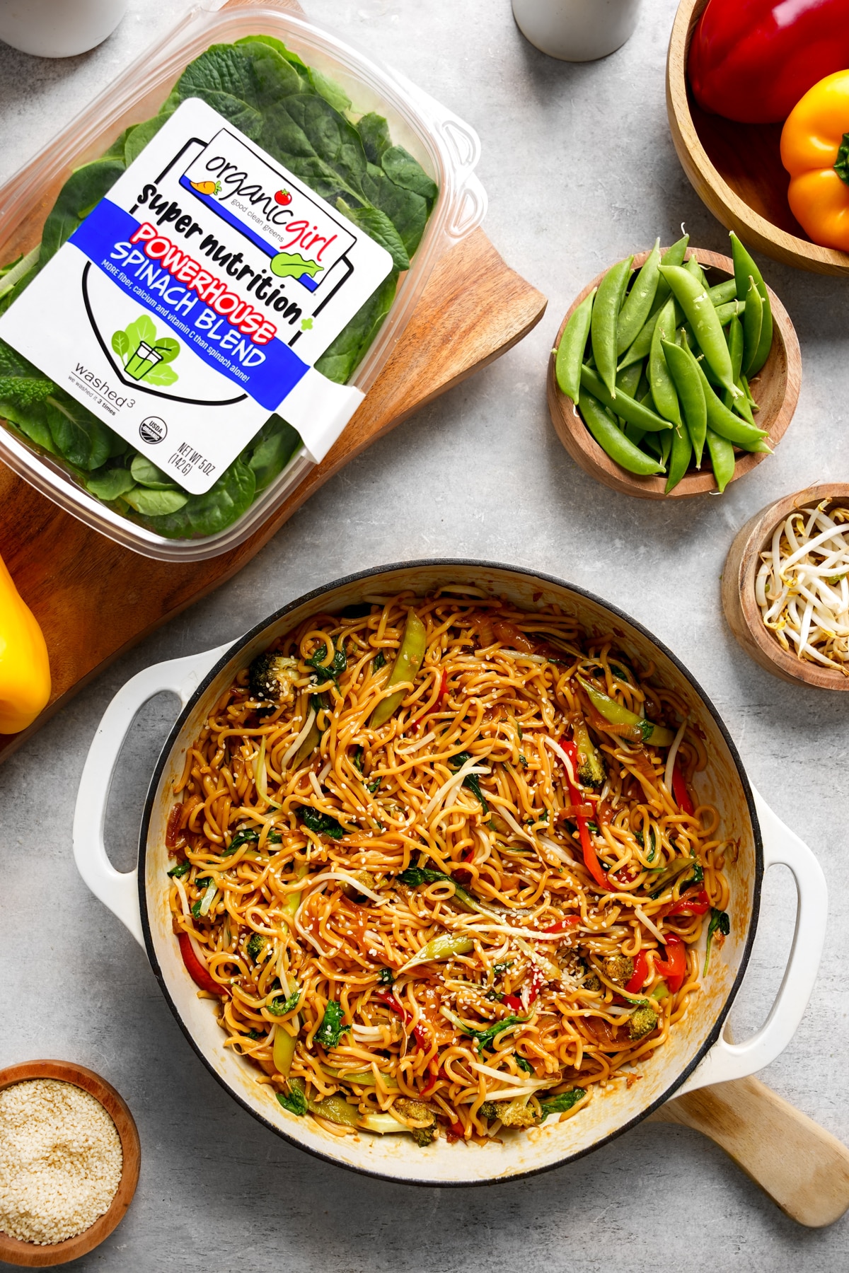 A pot of healthy vegan chow mein made with noodles, colorful vegetables, and sesame seeds, surrounded by fresh ingredients including Organicgirl Powerhouse Spinach Blend, snap peas, bean sprouts, and bell peppers. The setup showcases a wholesome plant-based meal with vibrant colors and fresh produce.