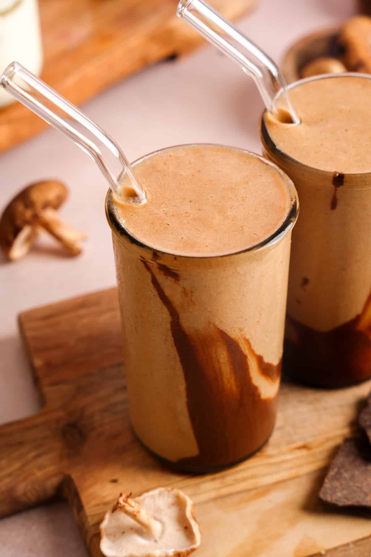 Two tall glasses of creamy tan-colored smoothie with rich chocolate swirls inside, served with clear glass straws on a wooden board. Shiitake mushrooms and pieces of dark chocolate are scattered around the board, suggesting the smoothie may be infused with mushroom or adaptogenic ingredients.