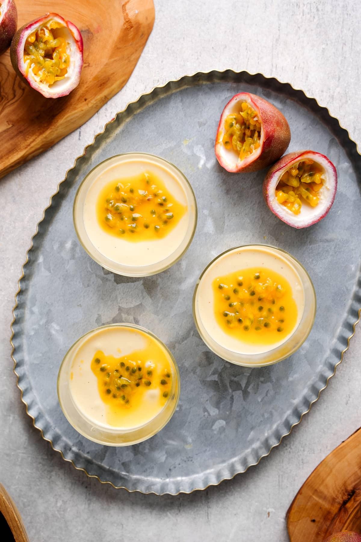Three glass cups filled with creamy vegan passion fruit mousse are arranged on a silver tray, each topped with bright golden passion fruit pulp and seeds. Around the tray, halved passion fruits with their vibrant interiors are displayed on wooden boards, adding a tropical touch to the elegant, plant-based dessert scene.