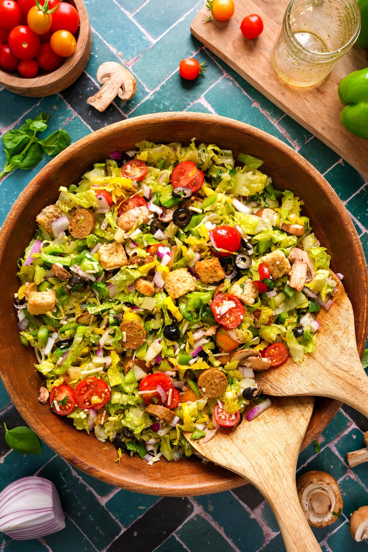 A vibrant chopped salad served in a large wooden bowl, filled with romaine lettuce, cherry tomatoes, sliced mushrooms, diced green bell peppers, red onions, black olives, banana peppers, vegan sausage slices, and golden croutons. The salad is tossed in a light vinaigrette and garnished with shredded vegan cheese. Wooden salad tongs rest inside the bowl, and the scene is surrounded by ingredients like cherry tomatoes, fresh basil, mushrooms, and a glass jar with remaining dressing on a teal-tiled surface.