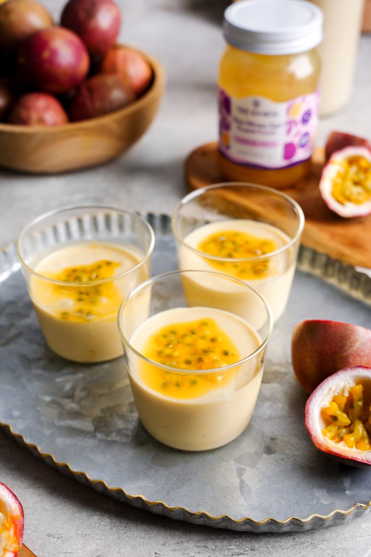 Three glass cups of creamy vegan passion fruit mousse topped with golden passion fruit pulp are arranged on a metal tray. In the background, a jar of True Sea Moss Passion Fruit Gel and a bowl of fresh passion fruits are visible, emphasizing the tropical and nutrient-rich ingredients used in this dairy-free dessert.