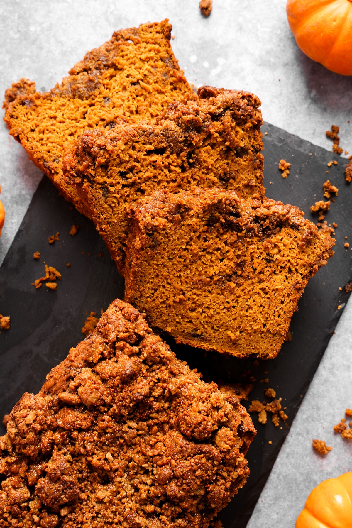 Close-up of moist vegan pumpkin crumb bread sliced on a black slate board. The golden-orange spelt loaf features a crispy, cinnamon-spiced crumble topping and is surrounded by mini pumpkins on a light gray background.