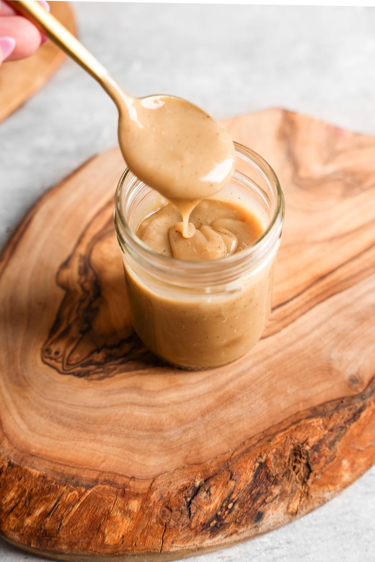 **Alt Text:**
A glass jar filled with creamy vegan sweetened condensed milk placed on a wooden board. A gold spoon is held above the jar, dripping the thick, smooth mixture back in, showcasing its rich and glossy texture.