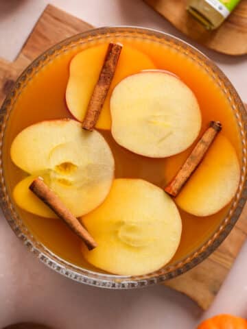 Overhead view of a glass punch bowl filled with spiced apple pumpkin cider topped with apple slices and cinnamon sticks, styled on a wooden board with autumn decor