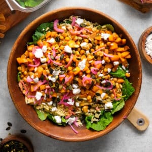 A vibrant vegan butternut squash and kale salad served in a wooden bowl, featuring roasted squash, chickpeas, orzo, pickled red onions, pumpkin seeds, and crumbled vegan feta over fresh baby kale. The colorful mix of ingredients creates a nourishing, hearty fall-inspired salad.