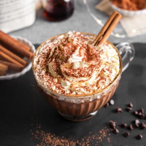Creamy vegan protein hot chocolate in a clear glass mug topped with whipped cream, cocoa powder, and cinnamon sticks, styled on a dark surface with chocolate chips and cozy holiday accents.