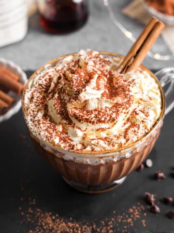 Creamy vegan protein hot chocolate in a clear glass mug topped with whipped cream, cocoa powder, and cinnamon sticks, styled on a dark surface with chocolate chips and cozy holiday accents.