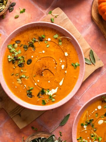 Overhead view of creamy vegan pumpkin soup in a pink bowl, garnished with pumpkin seeds, microgreens, coconut cream drizzle, and black pepper, set on a wooden board with garlic, sage leaves, and small pumpkins on a warm terracotta background.
