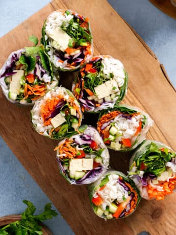 Fresh vegan tofu spring rolls sliced in half and arranged on a wooden cutting board, filled with rice noodles, tofu, shredded carrots, red cabbage, cucumber, microgreens, and herbs on a light blue background.