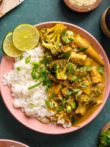 Bowl of vegan yellow curry served with fluffy white rice, tofu, broccoli, baby corn, and vegetables in a creamy coconut curry sauce, garnished with fresh cilantro, sesame seeds, and lime wedges.