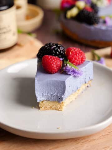 Close-up of a vegan butterfly pea flower cheesecake slice with a smooth purple filling and shortbread crust, topped with raspberries, blackberry, and edible flowers on a neutral plate.