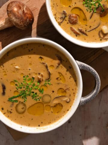 Two bowls of creamy vegan mushroom soup topped with fresh chives and olive oil, served with whole mushrooms on a wooden board — easy dairy-free mushroom soup recipe.
