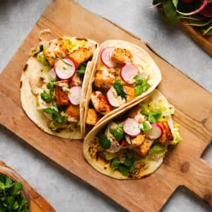 Three vegan tofu tacos on lightly charred tortillas filled with crispy tofu cubes, sliced radishes, shredded lettuce, cilantro, and a creamy drizzle, served on a rustic wooden cutting board.