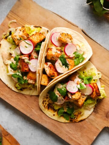 Three vegan tofu tacos on lightly charred tortillas filled with crispy tofu cubes, sliced radishes, shredded lettuce, cilantro, and a creamy drizzle, served on a rustic wooden cutting board.