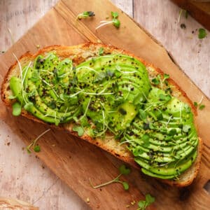Toasted slice of rustic bread topped with sliced avocado, microgreens, and sprinkled seasoning, served on a wooden board.