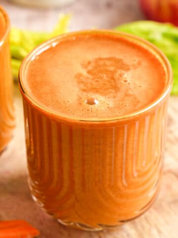 Fresh carrot celery juice served in a glass with a frothy top, surrounded by whole carrots, apples, celery, and a bottle of juice in the background.