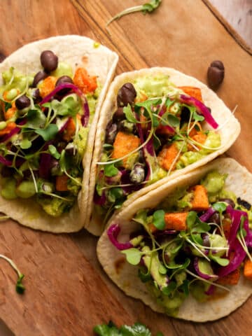 Three open-faced sweet potato tacos on small tortillas topped with guacamole, black beans, pickled red onions, microgreens, and creamy green dressing, arranged on a wooden cutting board with a bag of sweet potato croutons blurred in the background.
